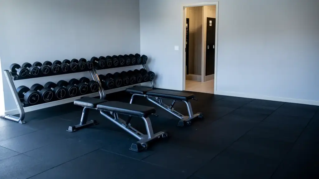Racked dumbbells, wiped padded benches and a black rubber floor with a locker room doorway beyond.